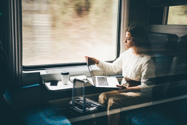 Kvinna sitter med laptop i tågkupé med resväska framför sig.